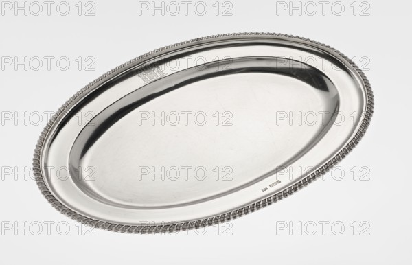 Oval platter, goldsmith/silversmith/dealer maker, purchase, scrap - purchase; features coats of arms, godron ornaments, rank crowns heraldry, earl's coronet heraldry. Oval platter with smooth mirror, slightly raised collar surrounded by a godron half-cylinder; on the collar a four-field coat of arms under an earl's coronet. 1904/1905. England; Sheffield, Great Britain. Dish. Height 2 cm; width 33 cm; depth 22.5 cm; weight 625 g., A polished oval metal tray with a raised rim and a twisted rope-like decorative edge, shown against a plain light background; the tray surface is shiny and reflective with visible light scratches and subtle tonal variations, displaying highlights and darker reflections in shades of silver, gray, and white.