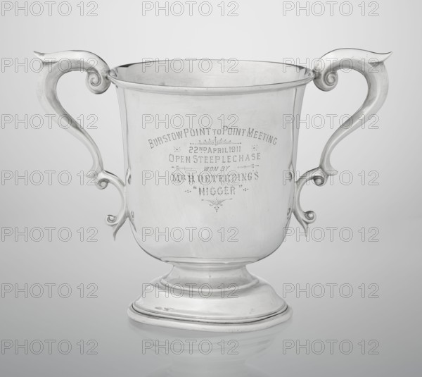 Prize cup by Mappin & Webb Ltd goldsmith, silversmith, dealer; fl. 1859-. Cup on a circular, arched, profiled base with a short shaft; semi-ovoid body flattened at the bottom; slightly flared upper edge bordered with a half-cylinder; two volute handles on the sides. Engraved inscription on the belly: BURSTON POINT TO POINT MEETING / 22nd APRIL 1911 / OPEN STEEP LECHASE / WON BY / MR H. DETERDING'S / 'NIGGER'. Dated 1910/1911, made in London, England. Vessel; height 16.5 cm, width 23.5 cm, depth 14.5 cm, weight 500 g., A silver trophy cup with a reflective metallic surface and ornate curved handles on each side, set against a light gray gradient background; the cup has a rounded body on a stepped circular base and visible engraved markings on its front; the overall colors are silver, light gray, and subtle darker gray reflections.