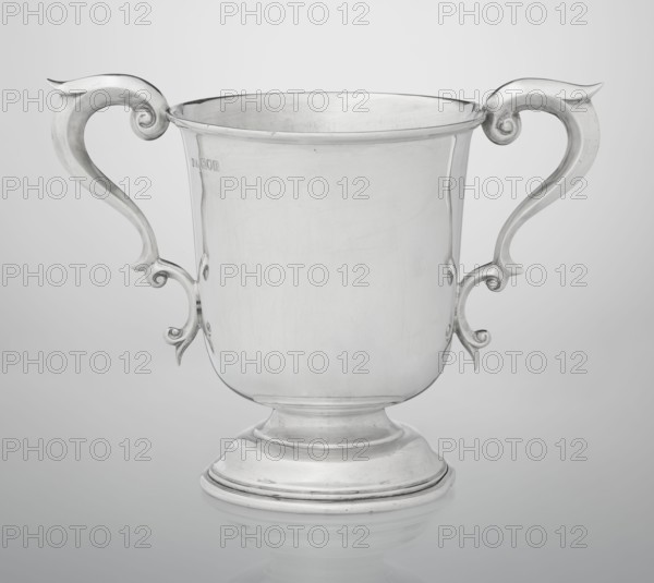 Prize cup by Mappin & Webb Ltd goldsmith, silversmith, dealer; fl. 1859-. Cup on a circular, arched, profiled base with a short shaft; semi-ovoid body flattened at the bottom; slightly flared upper edge bordered with a half-cylinder and two volute handles on the sides. Engraved on the belly: BURSTON POINT TO POINT MEETING / 22nd APRIL 1911 / OPEN STEEP LECHASE / WON BY / MR H.DETERDING'S / 'NIGGER'. Dated 1910/1911. England, London. Vessel; height 16.5 cm, width 23.5 cm, depth 14.5 cm, weight 500 g., A polished silver-colored metal trophy cup with a rounded footed base and a wide cylindrical body, shown front-facing against a light gray background; the cup has a pair of symmetrical ornate handles with curved scroll-like shapes extending outward and upward, and the surface of the metal shows subtle scratches and reflections.