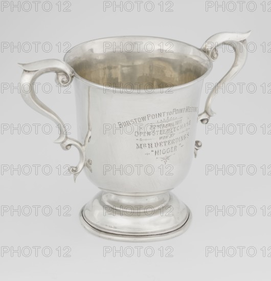 Prize cup by Mappin & Webb Ltd goldsmith, silversmith, dealer; fl. 1859-. Scrap - purchase, occasional inscriptions; sports award for horse racing cross-country steeplechase. Cup on a circular, arched, profiled base and short shaft; semi-ovoid body, flattened at the bottom; slightly flared upper edge bordered with a half-cylinder; two volute handles. Engraved inscription: BURSTON POINT TO POINT MEETING / 22nd APRIL 1911 / OPEN STEEP LECHASE / WON BY / MR H. DETERDING'S / 'NIGGER'. Dated 1910/1911. England, London. Vessel; height 16.5 cm, width 23.5 cm, depth 14.5 cm, weight 500 g., A polished silver trophy cup with a rounded rim and a flared pedestal base sits against a light gray background; it has ornate, curled handles on either side with decorative scrollwork and a smooth, reflective surface that shows subtle scratches and reflections, and the interior appears slightly darker and more golden-toned compared with the exterior; overall colors visible are silver, light gray, and a hint...