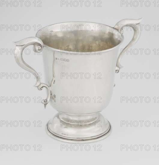 Prize cup, Mappin & Webb Ltd goldsmith, silversmith, dealer; fl. 1859- corporate maker; scrap - purchase, occasional inscriptions; sports award for horse racing cross-country steeplechase. Cup on a circular, arched, profiled base and a short shaft; semi-ovoid body, flattened at the bottom; slightly flared upper edge bordered with a half-cylinder, two volute handles on the sides. Engraved inscription: BURSTON POINT TO POINT MEETING / 22nd APRIL 1911 / OPEN STEEP LECHASE / WON BY / MR H.DETERDING'S / \NIGGER\, 1910/1911. England, London; vessel; height 16.5 cm, width 23.5 cm, depth 14.5 cm; weight 500 g., A polished silver cup with a rounded base and pedestal sits against a light gray background, featuring a flared rim and a slightly reflective interior showing muted golden and silver tones; the cup has a pair of ornate curved handles with scroll-like flourishes attached to opposite sides, and the exterior surface shows subtle scratches and a few small marks, with visible silver highligh...