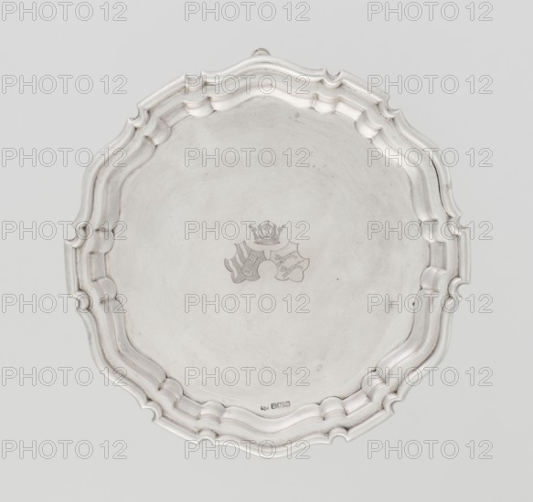 Tray on legs, goldsmith/silversmith, circular with three hoof-shaped legs; slightly raised, multi-partite profiled and ribbed collar with serrated edges. Two coats of arms engraved in the mirror beneath a noble crown. Inscription in italics on the bottom center: 29.9.1906 /von HvG. 1906/1907. Sheffield, England, Great Britain. Table accessory; height 2.4 cm, diameter 21.2 cm, weight 350 g., A round metal tray with a scalloped, layered rim and a smooth, slightly worn central surface, showing a small decorative emblem in the center; the tray appears in shades of silver and gray with subtle darker tarnish marks and light reflections.