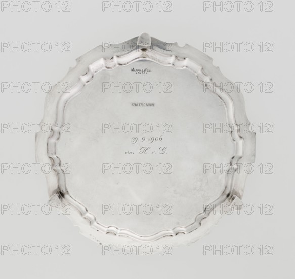 Tray on legs, maker: Mappin & Webb Ltd goldsmith, silversmith; fl. 1859–, circular silver tray supported on three hoof-shaped legs, slightly raised multi-partite profiled and ribbed collar with serrated edges; ribbed; two coats of arms engraved in the mirror under a noble crown; inscription in italics on the bottom center: 29.9.1906 /von HvG; dated 1906/1907; table accessory; height 2.4 cm, diameter 21.2 cm, weight 350 g; Sheffield, England., A round metallic tray with a scalloped, ornate rim and a slightly raised edge sits against a pale background; the tray appears silver in color with areas of darker tarnish and subtle surface scratches, and features a smooth central area surrounded by a decorative undulating border and a few small raised feet along the perimeter.