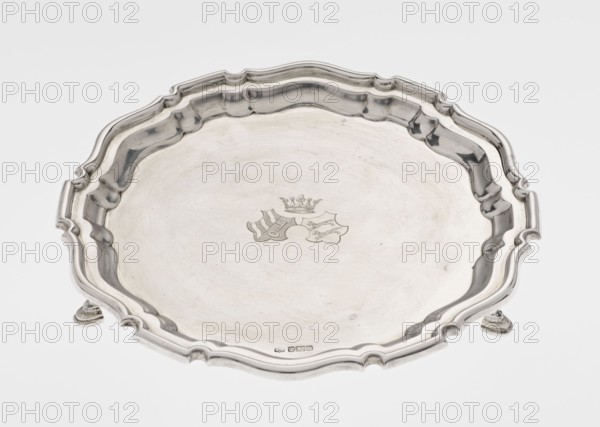 Tray on legs, goldsmiths Mappin & Webb Ltd maker, circular tray supported on three hoof-shaped legs, slightly raised multi-partite profiled and ribbed collar with serrated edges; ribbed surface. Two coats of arms engraved in the mirror under a noble crown. Inscription in italics on the bottom center: 29.9.1906 /von HvG. 1906/1907. Sheffield, England, Great Britain. Table accessory. Height 2.4 cm; diameter 21.2 cm; weight 350 g., A shallow round tray with a scalloped, ornate rim and raised edge resting on small curved feet, shown from above; the tray appears metallic with a reflective silver surface showing subtle scratches and faint wear; a decorative engraved emblem is visible near the center; the rim and inner contours create shadows in darker silver and gray tones against a white background, and the overall colors visible are silver, gray, and white.