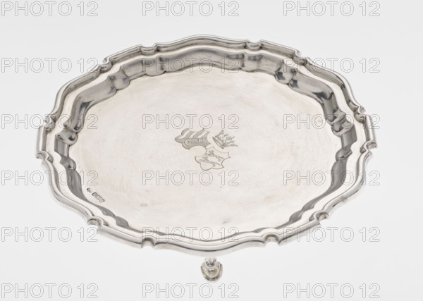 Tray on legs, Mappin & Webb Ltd goldsmith, silversmith, dealers; fl. 1859-, maker, scrap - purchase, coats of arms, rank crown heraldry, ribbed; circular tray supported on three hoof-shaped legs, with a slightly raised, multi-partite, profiled and ribbed collar with serrated edges. Two coats of arms are engraved in the mirror under a noble crown. On the bottom in the middle, an inscription in italics is engraved: 29.9.1906 /von HvG. Dated 1906/1907. Table accessory; height 2.4 cm, diameter 21.2 cm, weight 350 g., A silver tray with a scalloped, ornate rim sits against a white background, showing a shallow, slightly convex surface with a subtle central engraved motif and faint surface scratches; the rim has curved, raised borders and recessed sections creating a decorative pattern, and a small portion of a matching silver foot is visible at the bottom edge. Colors visible include silver tones and white.