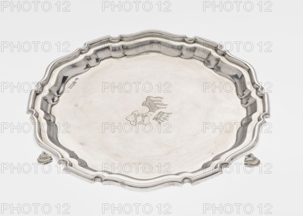 Tray on legs, goldsmith/silversmith maker: Mappin & Webb Ltd. Circular tray supported on three hoof-shaped legs, with a slightly raised, multi-partite profiled and ribbed collar with serrated edges. Ribbed surface; two coats of arms engraved in the mirror beneath a noble crown. Inscription in italics on the bottom center: 29.9.1906 /von HvG. Date: 1906/1907. Material: presumably silver table accessory. Dimensions: height 2.4 cm, diameter 21.2 cm, weight 350 g. Location made: Sheffield, England., A shallow decorative metal tray with a scalloped, ornate rim and a slightly raised center, shown from above; the tray has a smooth reflective surface with subtle scratches and a faint engraved emblem near the center; the rim has molded curves and a few raised sections and stands on small feet that are partly visible; the visible colors are silver tones, gray, and off-white from reflections.