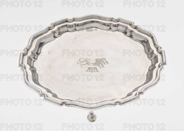 Tray on legs, ribbed circular tray supported on three hoof-shaped legs, with a slightly raised, multi-partite, profiled and ribbed collar with serrated edges; two coats of arms engraved in the mirror under a noble crown. Inscribed in italics on the bottom: 29.9.1906 / von HvG. Maker: Mappin & Webb Ltd goldsmith, silversmith, dealers, 1906/1907. Material/type: table accessory. Dimensions: height 2.4 cm, diameter 21.2 cm, weight 350 g., A round metal tray with a scalloped raised rim and a decorative molded edge, shown from above on a pale background; the tray surface is smooth with some subtle scratches and a small engraved decorative motif near the center; the metal is silvery with reflective highlights and muted gray shadows.