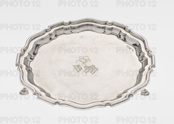 Tray on legs, Mappin & Webb Ltd goldsmith, silversmith, dealers; fl. 1859–, maker, scrap - purchase, coats of arms, rank crown heraldry, ribbed; circular tray supported on three hoof-shaped legs, with a slightly raised, multi-partite, profiled and ribbed collar with serrated edges. Two coats of arms are engraved in the mirror under a noble crown. On the bottom in the middle, an inscription in italics is engraved: 29.9.1906 /von HvG. Dated 1906/1907. Table accessory; height 2.4 cm, diameter 21.2 cm, weight 350 g; made in Sheffield, England., A shallow round metal tray with a scalloped, raised rim and ornate curved edge sits on small rounded feet, showing a reflective surface with a subtle central decorative engraved motif; the tray appears in shades of silver and gray against a pale off-white background.