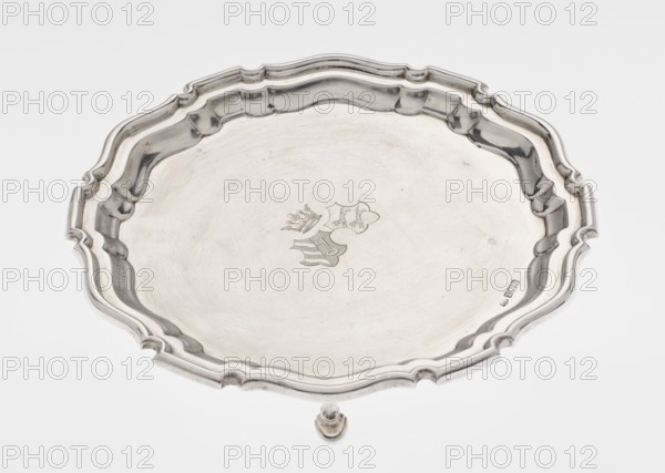 Tray on legs, goldsmith/silversmith/ dealer: Mappin & Webb Ltd maker, scrap - purchase, coats of arms, rank crown heraldry, ribbed; circular tray supported on three hoof-shaped legs, with a slightly raised, multi-partite, profiled and ribbed collar with serrated edges. Two coats of arms engraved in the mirror beneath a noble crown. Inscription in italics on the bottom center: 29.9.1906 /von HvG. Dated 1906/1907. Sheffield, England. Table accessory. Height 2.4 cm; diameter 21.2 cm; weight 350 g., A decorative round metal tray with a scalloped, raised edge and an ornate rim, shown from above; the tray has a smooth central surface with a faint engraved emblem and a small raised foot visible at the bottom edge. The visible colors are silver, light gray, and subtle darker gray reflections.