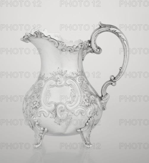 Milk jug by Mappin & Webb Ltd goldsmith, silversmith, dealer; fl. 1859–, neo-rococo style, 1895/1896. Pear-shaped milk jug supported on four volute legs, decorated with an acanthus leaf at the base; belly decorated with rococo motifs in the form of channel-shaped jars, rocailles, flowers, and acanthus leaves; volute handle with acanthus projections; wide raised spout with serrated edge. Material: presumed silver. Dimensions: height 14.4 cm, width 9.5 cm, depth 8.5 cm, weight 266 g. Made in Sheffield, England., A metallic decorative pitcher with an ornate handle and scalloped rim sits on a reflective surface; the pitcher has embossed floral and scrollwork patterns on its bulbous body and stands on small curved feet. The visible colors are shades of silver and gray with subtle highlights and shadows.