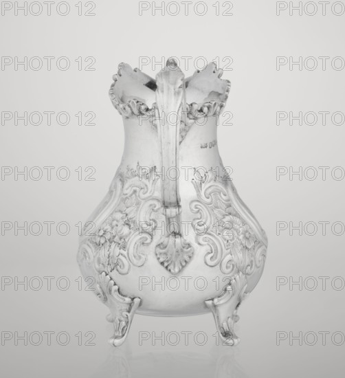 Milk jug by Mappin & Webb Ltd goldsmith/silversmith, neo-rococo style, 1895–1896. Pear-shaped milk jug supported on four volute legs, decorated with an acanthus leaf at the base; belly ornamented with rococo motifs including channel-shaped jars, rocailles, flowers and acanthus leaves. Volute handle with acanthus projections and a wide raised spout with serrated edge. Material: metal. Place: Sheffield, England. Function: vessel. Dimensions: height 14.4 cm, width 9.5 cm, depth 8.5 cm; weight 266 g., A silver-colored ornate pitcher with a scalloped rim and a single curved handle, decorated with raised floral and scrollwork patterns on its bulbous body and standing on a few decorative feet, shown against a light gray background.
