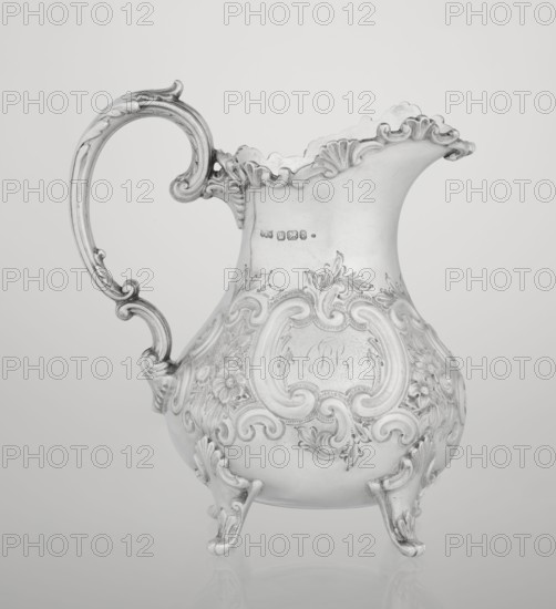Milk jug by Mappin & Webb Ltd goldsmith, silversmith, dealer; fl. 1859–, neo-rococo style, pear-shaped milk jug supported on four volute legs, decorated with an acanthus leaf at the base and on the belly with rococo motifs in the form of channel-shaped jars, rocailles, flowers and acanthus leaves; volute handle with acanthus projections and a wide, raised spout with a serrated edge; 1895–1896; Sheffield, England, Great Britain; vessel; height 14.4 cm, width 9.5 cm, depth 8.5 cm; weight 266 g., A single ornate metal pitcher with a curved handle and decorative scalloped rim sits against a plain background; the surface shows elaborate raised floral and scrollwork patterns across its rounded body and feet, and the handle features curled, carved details; the image appears in shades of gray, silver, and white.