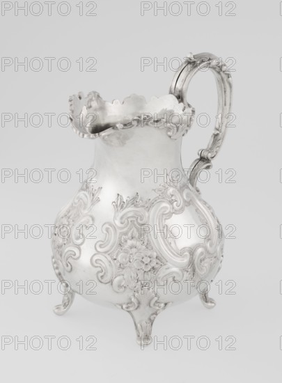 Milk jug by Mappin & Webb Ltd goldsmith, silversmith, dealer; fl. 1859–, neo-rococo style, pear-shaped milk jug supported on four volute legs, decorated with an acanthus leaf at the base and on the belly with rococo motifs in the form of channel-shaped jars, rocailles, flowers and acanthus leaves; volute handle with acanthus projections and a wide, raised spout with a serrated edge; dated 1895/1896; Sheffield, England, Great Britain; vessel; height 14.4 cm, width 9.5 cm, depth 8.5 cm; weight 266 g., A decorative metal pitcher with an ornate curved handle and a scalloped rim sits against a pale background; the pitcher has a bulbous body raised on a few molded feet and is covered in raised floral and scrollwork ornamentation, showing a shiny metallic surface in tones of silver and gray.