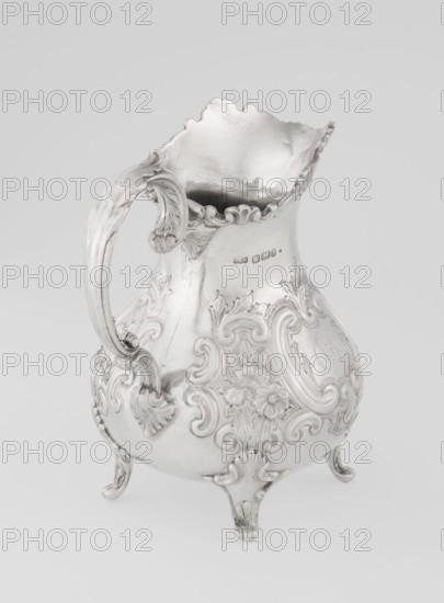 Milk jug by Mappin & Webb Ltd goldsmith, silversmith, dealer; fl. 1859–, neo-rococo style, 1895–1896. Pear-shaped milk jug supported on four volute legs, decorated with an acanthus leaf at the base; belly decorated with rococo motifs in the form of channel-shaped jars, rocailles, flowers and acanthus leaves; volute handle with acanthus projections and a wide raised spout with a serrated edge. Materials/technique: metal silver; function: vessel. Dimensions: height 14.4 cm, width 9.5 cm, depth 8.5 cm; weight 266 g. Made in Sheffield, England., A silver pitcher with an ornate curved handle and a scalloped, flared rim sits on a plain light background; the pitcher has an embossed floral and scrollwork design covering its bulbous body, rests on three decorative feet, and shows a few reflective highlights and subtle shadows in tones of silver and gray.