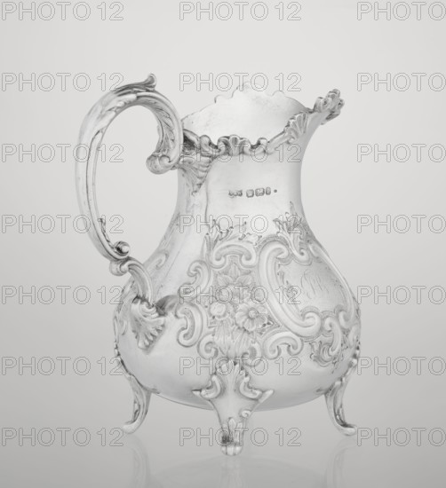 Milk jug by Mappin & Webb Ltd goldsmith, silversmith, dealer; fl. 1859–, neo-rococo style, 1895–1896. Pear-shaped milk jug supported on four volute legs, decorated with an acanthus leaf at the base; belly decorated with rococo motifs in the form of channel-shaped jars, rocailles, flowers, and acanthus leaves; volute handle with acanthus projections; wide raised spout with serrated edge. Material: metal silver. Sheffield, England. Vessel; height 14.4 cm, width 9.5 cm, depth 8.5 cm, weight 266 g., A decorative metal pitcher with an ornate curved handle and a scalloped rim stands on a reflective surface; the pitcher features embossed floral and scrollwork patterns across its bulbous body and rests on three stylized feet, showing shades of silver, gray, and white.