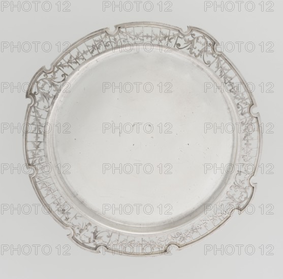 Round tray, circular silver tray with smooth mirror, slightly raised, openwork collar decorated with plant tendril, irregularly cut edge bordered with profiled band, maker: Mappin & Webb Ltd goldsmith, silversmith, dealer; fl. 1859-, date: 1922/1923, place: Sheffield, England, Great Britain; function: table accessory; dimensions: height 1.8 cm, width 27.1 cm, depth 18 cm; weight: 576.5 g., A round metal tray with a smooth central surface and an ornate pierced rim featuring decorative scrollwork and floral motifs; the tray and its rim are shades of silver and light gray, set against a pale gray background.