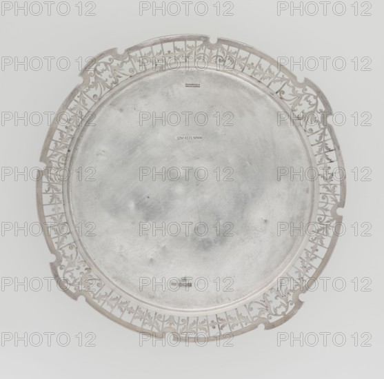 Round tray, goldsmith/silversmith Mappin & Webb Ltd fl. 1859-; circular silver tray with smooth mirror, slightly raised with openwork collar decorated with plant tendril; irregularly cut edge bordered with profiled band; openwork; 1922/1923; Sheffield, England; table accessory. Height 1.8 cm; diameter 27.1 cm; depth 18 cm; weight 576.5 g., A round metallic tray with a raised rim and an openwork decorative border featuring floral and scroll motifs, the tray surface showing a worn, brushed texture with small dents and scuffs; the visible colors are silver and gray with subtle variations in tone.