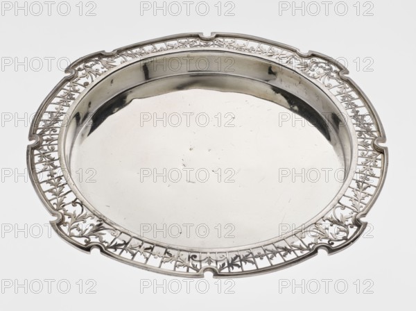 Round tray; maker: Mappin & Webb Ltd goldsmith, silversmith, dealer; fl. 1859-; function: table accessory. Circular silver tray with smooth mirror, slightly raised, openwork collar decorated with plant tendrils, irregularly cut edge bordered with profiled band. Inscription/marking: maker's name. Date: 1922/1923. Place: Sheffield, England. Dimensions: height 1.8 cm; width 27.1 cm; depth 18 cm. Weight: 576.5 g., A round metal tray with a reflective surface and an ornate pierced rim featuring a repeating floral and lattice pattern, shown against a plain light background; the tray is silver in color with areas of darker reflection and slight surface scratches.