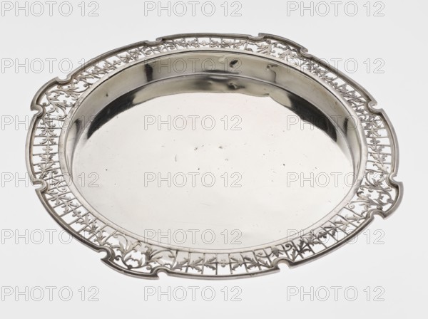 Round tray, circular silver with smooth mirror and slightly raised, openwork collar decorated with plant tendril, irregularly cut edge bordered with profiled band; maker: Mappin & Webb Ltd goldsmith, silversmith, dealer; fl. 1859-; date: 1922/1923; place: Sheffield, England; function: table accessory; dimensions: height 1.8 cm, width 27.1 cm, depth 18 cm; weight: 576.5 g., A round metal tray with a reflective central surface and an ornate pierced rim is shown, primarily silver in color with highlights and darker reflections, set against a light gray background; the rim features an intricate openwork pattern with flowing, curving shapes and a few scalloped notches around the edge, while the central area is smooth and shiny with a few small surface marks.