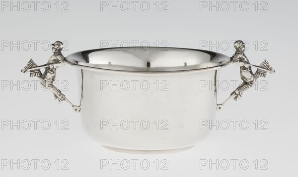 Porridge bowl porringer, by Mappin & Webb Ltd goldsmith/silversmith, fl. 1859–. Cylindrical bowl with flat bottom, slightly widening toward the top with a turned-up edge edged by a smooth half-cylinder. Two side handles modeled as figurines of boys dressed in jockey outfits, riding toy horses on sticks. Associated with porridge/porringer, children's games. Dated 1935–1936. England Sheffield. Vessel; height 5.6 cm, width 16.3 cm, depth 10.5 cm, weight 125 g., A polished metal bowl with a rounded body and flared rim sits against a pale background, showing reflective surfaces and highlights. Attached at opposite sides of the rim are sculpted metal handles in the form of riders on stylized horses, each figure extending outward from the bowl. The bowl and handles display metallic silver tones with areas of darker reflection and subtle shadowing, and the background is a gradient of light gray to off-white.