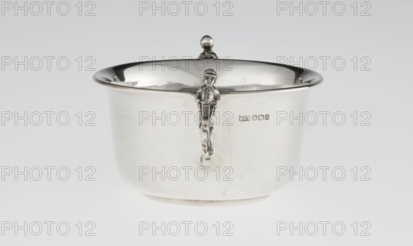 Porridge bowl - porringer. Maker: Mappin & Webb Ltd goldsmith, silversmith, dealer; fl. 1859-. Cylindrical bowl with a flat bottom, slightly widening towards the top, with a turned-up edge edged with a smooth half-cylinder. Two handles on the sides in the shape of figurines of boys dressed in jockey outfits, riding toy horses on sticks. Function: porridge dish/porringer; also associated with children's games, toy horses. Date: 1935/1936. Place: Sheffield, England, Great Britain. Dimensions: height 5.6 cm; width 16.3 cm; depth 10.5 cm; weight 125 g., A shiny metal bowl with a rounded rim sits against a pale background, showing a reflective interior surface and a smooth exterior. The bowl has a small decorative handle or figure attached to its front edge that resembles a standing person, and a rounded finial or knob centered on the rim at the back. The visible colors are silver tones, light gray, and subtle darker gray reflections, with a mostly white to very light gray background.