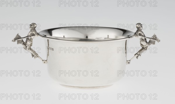 Porridge bowl porringer, maker: Mappin & Webb Ltd goldsmith, silversmith, dealer; fl. 1859-. Cylindrical bowl with flat bottom, slightly widening toward the top, with a turned-up edge edged by a smooth half-cylinder. Two side handles in the shape of figurines of boys dressed in jockey outfits, riding toy horses on sticks. Function: porridge dish/toy., vessel. Date: 1935/1936. Place: Sheffield, England, Great Britain. Dimensions: height 5.6 cm; width 16.3 cm; depth 10.5 cm; weight 125 g. Subjects/inscriptions: jockeys, children's games, porridge., A reflective metal bowl with a smooth rounded body and flared rim sits against a light gray background, featuring a pair of decorative handles shaped like riders on hobby horses extending horizontally from opposite sides; the bowl and handles are silver in color with darker gray reflections, and the background shows shades of pale gray and off-white.