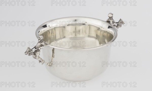Porridge bowl porringer, Mappin & Webb Ltd goldsmith, silversmith, dealer; fl. 1859-. Cylindrical bowl with a flat bottom, slightly widening toward the top, with a turned-up edge edged with a smooth half-cylinder. Two side handles in the form of figurines of boys dressed in jockey outfits, riding toy horses on sticks. Inscription/date: 1935/1936. Place: Sheffield, England, Great Britain. Function: vessel porridge dish. Dimensions: height 5.6 cm, width 16.3 cm, depth 10.5 cm; weight 125 g. Subjects/ motifs: jockeys, children's games, porridge., A shiny metal bowl with a reflective interior and exterior sits on a light gray surface, featuring a group of small sculpted figures attached to the rim on opposite sides that resemble riders with horses; the metal appears silver with highlights and soft shadows, and there are subtle reflections of beige and darker gray tones inside the bowl.
