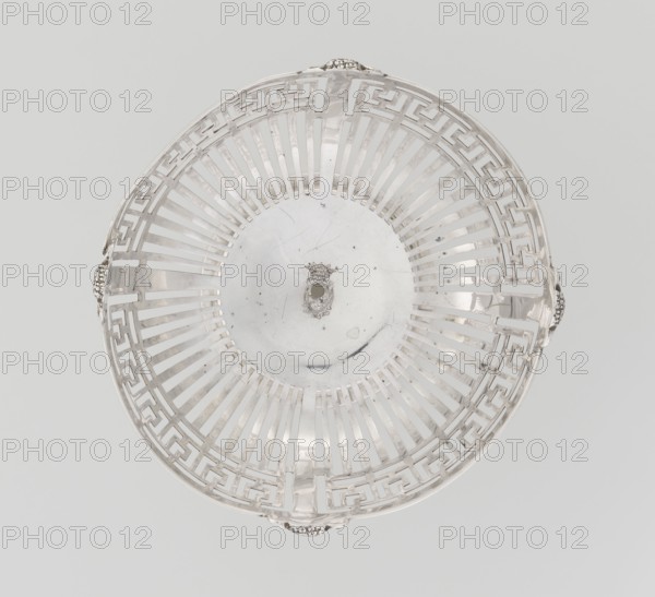 Openwork bowl by Mappin & Webb Ltd goldsmith, silversmith, sellers; fl. 1859-. Bowl on a circular, stepped foot and short shaft; hemispherical, decorated with radial openwork and a wide strip of stylised meander under the upper edge, enclosed in a smooth band; decorated with four relief ram's heads. At the bottom, a four-field coat of arms in an oval shield under an earl's count's crown. Inscribed/dated 1908/1909. England, Birmingham. Vessel. Materials not stated. Height 9 cm; diameter bowl rim 13.5 cm; weight 126 g., A round metal basket viewed from above with an open lattice pattern of radial and geometric cutouts, featuring ornate handles at the rim; the surface shows slight scratches and spots. The visible colors are silver, light gray, and a pale off-white background.