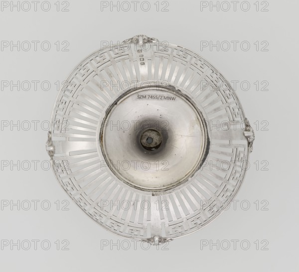 Openwork bowl by Mappin & Webb Ltd goldsmith, silversmith, sellers; fl. 1859-. Bowl on a circular stepped foot and short shaft, hemispherical, decorated with radial openwork and a wide strip of stylised meander under the upper edge, enclosed in a smooth band, and decorated with four relief ram's heads. At the bottom, a four-field coat of arms in an oval shield beneath an earl's count's crown. Inscription/markings reference 1908/1909. Material: metal. Function: vessel. Style: openwork, decorative ornamentation. Place: Birmingham, England, Great Britain. Dimensions: height 9 cm; rim diameter 13.5 cm; weight 126 g., A round metallic object with a central concave depression surrounded by a lattice-like openwork rim is shown from above, with a smooth inner bowl that has some surface marks and a dark circular opening at its center; the rim features repeating geometric cutouts and small ornamental protrusions at intervals, and the visible colors are silver, gray, and darker gray tones.