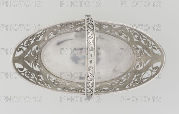 Bread Basket; maker: Mau, Hainrich fl. early 20th century maker; Hutton & Sons Ltd Sheffield, Birmingham, London; goldsmith and silversmith firm; fl. 1800–1930 maker; purchased provenance, scrap - purchased. Geometric and floral ornament; openwork; godron ornaments. Basket on four curved legs; oval, boat-shaped; sides decorated with openwork plant and floral twig motifs inscribed in an arched ogee back; ribbon reserves; ribbon upper edge; enclosed at the bottom by a godron half-cylinder. Handle in the form of a movable bow, openwork, decorated with plant tendrils. Date: 1901/1902, London, England, Great Britain. Vessel. Height 18 cm; width 22.7 cm; depth 3.7 cm; weight 388 g., An oval metal tray with a central plain recessed surface and an ornate pierced rim featuring repeating floral and scroll patterns, shown from above with a curved decorative handle crossing the center; the visible colors are silver, light gray, and subtle darker gray tones.