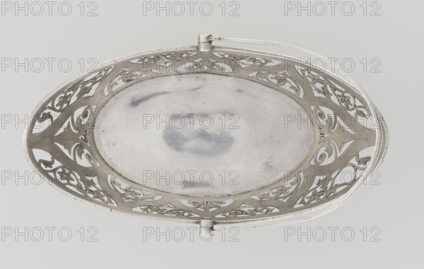 Bread basket by Hainrich Mau fl. early 20th century and Hutton & Sons Ltd goldsmith and silversmith firm, fl. 1800–1930. Oval, boat-shaped openwork silver basket on four curved legs, decorated on the sides with openwork plant and floral twig motifs set in an arched ogee back, ribbon reserves, ribbon upper edge, enclosed at the bottom by a godron half-cylinder. Handle is a movable bow in openwork decorated with plant tendrils. Geometric and floral ornament, godron ornaments. Dated 1901–1902, London, England, Great Britain. Vessel; height 18 cm, width 22.7 cm, depth 3.7 cm, weight 388 g., A decorative oval metal dish viewed from above with an openwork pierced border featuring ornate scroll and floral cutouts and a smooth central well showing subtle surface marks; the dish and its handles are metallic silver with areas of slightly darker gray patina against a pale gray background.