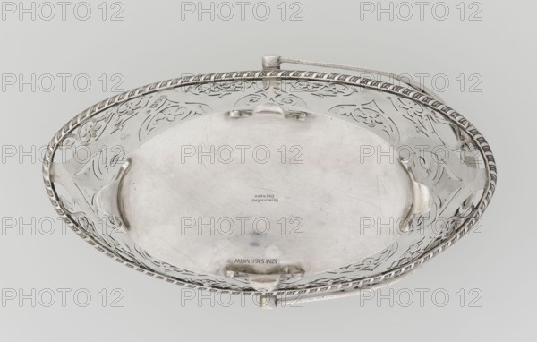 Bread basket by Mau, Hainrich fl. early 20th century and Hutton & Sons Ltd goldsmith and silversmith firm; fl. 1800–1930. Geometric and floral ornament; openwork; godron ornaments. Oval, boat-shaped basket on four curved legs, sides decorated with openwork plant and floral twig motifs inscribed in an arched ogee back, ribbon reserves, ribbon upper edge, enclosed at the bottom by a godron half-cylinder. Handle in the form of a movable bow, openwork, decorated with plant tendrils. Dated 1901/1902, London, England. Vessel; height 18 cm, width 22.7 cm, depth 3.7 cm, weight 388 g., An oval decorative metal dish with a raised ornate rim and pierced pattern around its edge, showing a smooth central basin with some surface scratches and small dark spots; the rim has a twisted rope-like border and floral or scroll cutout motifs, and there are a few small protruding tabs and a hinged handle attached at one side; visible colors include silver tones, pale gray, and subtle darker gray accents.
