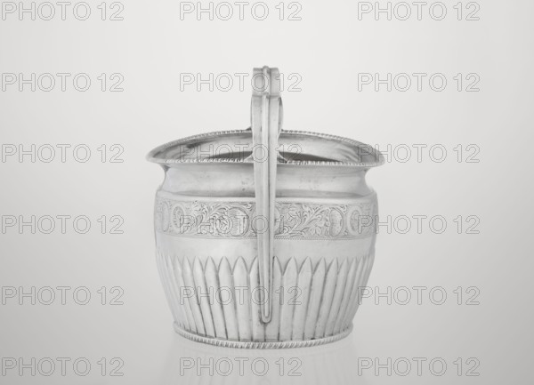 Sugar bowl by Hutton & Sons Ltd Sheffield, Birmingham, London; goldsmith and silversmith firm; fl. 1800–1930. Silver sugar bowl, oval with two ribbon handles; clasp vertically in the lower section; decorated above with a band of stylised acanthus twig; upper edge decorated with a godron half-cylinder. Godron ornaments, plant twig motif. Dated 1895–1896, London, England. Vessel sugar, height 11 cm, width 17.3 cm, depth 10.5 cm, weight 231 g., A metallic bucket-like container with a single vertical handle attached at the center, shown from the front. The container has decorative patterns around its upper band and a fluted lower section, with a ridged rim at the top and bottom. The surface appears smooth and reflective with subtle tarnish or patina, and the overall colors visible are shades of silver and gray against a light gray background.