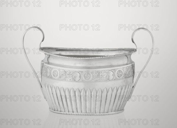 Sugar bowl by Hutton & Sons Ltd Sheffield, Birmingham, London; goldsmith and silversmith firm; fl. 1800–1930. Silver sugar bowl, oval with two ribbon handles; clasp vertically in the lower section; decorated above with a band of stylised acanthus twig; upper edge decorated with a godron half-cylinder. Godron ornaments, plant/acanthus twig motif. Date: 1895/1896, London, England, Great Britain. Function: sugar vessel. Dimensions: height 11 cm, width 17.3 cm, depth 10.5 cm; weight 231 g., A silver vessel with two upward-curving handles is centered against a light gray background; the vessel has a rounded rim with a beaded edge, an upper band decorated with an engraved floral and circular motif, and a lower section with vertical fluted ridges; the surface shows reflective highlights and subtle shadows, and the visible colors are silver, gray, and white.
