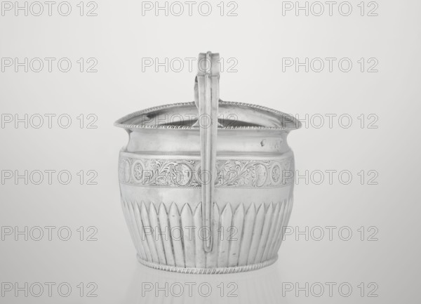 Sugar bowl by Hutton & Sons Ltd goldsmith and silversmith firm, active ca. 1800–1930. Silver sugar bowl, oval with two ribbon handles; clasp vertically in the lower section; decorated above with a band of stylized acanthus twig and an upper edge with a godron half-cylinder. Features godron ornaments and a plant twig motif. Dated 1895–1896, London, England. Vessel; height 11 cm, width 17.3 cm, depth 10.5 cm; weight 231 g., A metal vessel with a curved rim and a central vertical handle dominates the image; the surface shows ornate engraved patterns around the upper body and fluted vertical grooves along the lower body. The handle rises above the rim and has a simple rounded profile. The vessel rests on a flat reflective surface, and the background is a smooth, light gradient. Visible colors include shades of silver, gray, and off-white.