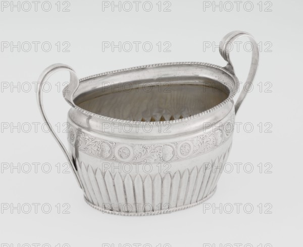 Sugar bowl by Hutton & Sons Ltd Sheffield, Birmingham, London; goldsmith and silversmith firm; active 1800–1930. Silver sugar bowl, oval with two ribbon handles; clasp vertically in the lower section. Decorated above with a band of stylised acanthus twig and an upper edge with a godron half-cylinder. Godron ornaments, plant twig motif. Function: sugar vessel. Inscription/markings: maker. Dated 1895–1896, London, England. Dimensions: height 11 cm, width 17.3 cm, depth 10.5 cm; weight 231 g., A metallic vessel with a silvery surface and a slightly reflective interior sits against a pale background; it has an oval opening, a decorative rim with a beaded edge, two tall curved handles rising from opposite sides, an engraved band of ornate swirling floral and circular motifs around its upper body, and a lower section with vertical fluted ridges, showing shades of silver, gray, and subtle warm reflections inside.