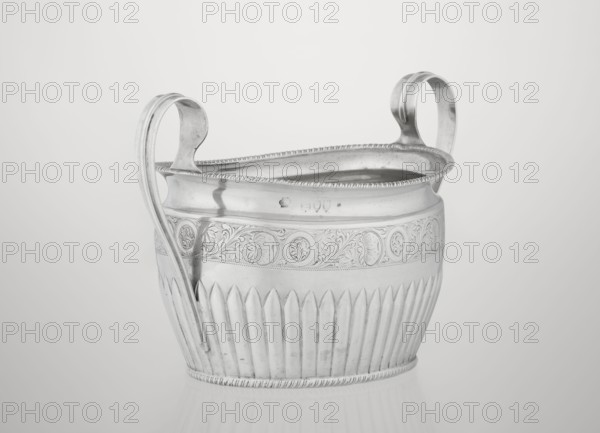 Sugar bowl by Hutton & Sons Ltd goldsmith and silversmith firm; active c. 1800–1930. Silver sugar bowl, oval with two ribbon handles and a clasp vertically in the lower section. Decorated above with a band of stylised acanthus twig; upper edge decorated with a godron half-cylinder. Features godron ornaments and a plant/acanthus twig motif. Dated 1895/1896, London, England. Vessel: height 11 cm, width 17.3 cm, depth 10.5 cm, weight 231 g., A metallic vessel with a reflective silver surface, featuring two curved handles that rise above its rim, sits against a light gray background. The vessel has a decorative band near the top with embossed floral and circular motifs, a beaded rim, and vertical fluted panels along its lower half. The handles have smooth, rounded contours and the metal shows subtle highlights and shadows that emphasize its shape and texture. The overall colors visible are silver and shades of gray.