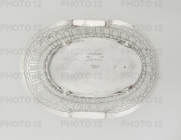 Fruit Basket, Mau, Hainrich fl. early 20th century maker; Hutton & Sons Ltd Sheffield, Birmingham, London; goldsmith and silversmith firm; fl. 1800-1930 maker, scrap - purchase, openwork, neoclassicism style, geometric ornaments, garlands ornament, pearling ornament, neoclassical, openwork, oval basket, supported on four volute legs, openwork formed by bands of ovals, circles and rectangles; top and bottom edges bordered with pearling; raised sides topped on the shorter axis semi-plastic vase with laurel leaf garlands. Engraved on the bottom with an italic inscription: 6. September / 1893 1918 / von CP Dietz, 1908/1909. England, London, Great Britain. Vessel, height 8.5 cm, width 35.5 cm, depth 25 cm, weight 925 g, A metallic oval tray with an open lattice rim featuring geometric and circular cutouts, shown from above on a pale gray background; the tray has a central smooth recessed area with faint surface scratches and small dark blemishes, and a few raised supports along the inner ed...