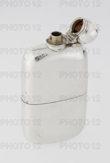 Hip flask, Unite, George & Sons Birmingham; goldsmith's firm; fl. 1825–1928 maker; so-called cuboid hip flask with rounded corners; smooth, slightly curved; spout in the form of a narrow neck with an applied spherical hinged stopper; bottle narrowed in the lower part from the middle; on this part a slide-in container — a cup masking the narrowing is placed; the interior of the cup and the spout are gilded; 1907/1908; England, Birmingham, Great Britain; vessel; height 15.5 cm, width 7.6 cm, depth 2.7 cm; weight 240 g., A smooth metallic object with a polished silver surface and rounded edges is shown against a light gray background, featuring an open circular cap revealing an inner cavity with a brown interior, a short cylindrical neck with a slightly tarnished brass-colored rim, and a hinge connecting the cap to the body; the surface shows subtle scratches and small dark blemishes, and there are a few small raised marks on the upper area near the neck.