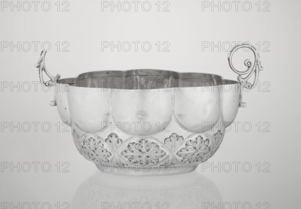 Porridge bowl porringer, Wakely & Wheeler Ltd London; goldsmiths; fl. 1791–1958, hemispherical bowl decorated with relief acanthus rosettes in the lower part, knobbed above, octagonal-shaped rim; two volute handles on the sides one half-preserved. Materials/technique: silver implied. Function: porridge dish. Ornament: rosette, acanthus, relief decoration. Date: 1891/1892. Place: London, England, Great Britain. Dimensions: height 7.6 cm; width 24 cm; depth 19.5 cm; weight 415 g., A decorative metal bowl with ornate handles sits centered on a smooth surface, showing a reflective finish in tones of silver and gray; the bowl has a scalloped rim and a rounded body with embossed floral-like patterns along the lower portion, and a subtle reflection of the bowl appears on the surface beneath it.