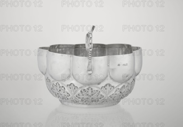 Porridge bowl porringer, Wakely & Wheeler Ltd London; goldsmiths; fl. 1791–1958 maker; hemispherical bowl decorated with relief acanthus rosettes in the lower part, knobbed above, octave-shaped rim; two volute handles on the sides one half-preserved; rosette ornament; relief decoration; porridge dish; 1891–1892; London, England, Great Britain; dish; height 7.6 cm, width 24 cm, depth 19.5 cm, weight 415 g, A silver bowl with a scalloped rim and a curved handle rising from its center, showing a reflective metallic surface in shades of silver and gray; the lower portion features a repeating floral cutout pattern around the body, and the inside shows darker gray reflections while the exterior displays lighter silver highlights and subtle scratches.