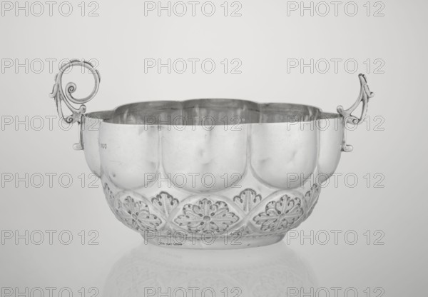 Porridge bowl porringer, Wakely & Wheeler Ltd London; goldsmiths; fl. 1791–1958 maker; porringer porridge dish with hemispherical bowl decorated with relief acanthus rosettes in the lower part, knobbed above, octave-shaped rim; two volute handles on the sides one half-preserved; rosette ornament; relief decoration; acanthus motif; 1891/1892; England, London, Great Britain; dish; height 7.6 cm, width 24 cm, depth 19.5 cm, weight 415 g., A metal bowl with a scalloped rim and ornate curved handles sits centered against a light neutral background, showing a reflective surface with highlights and subtle scratches; the bowl has a rounded body with embossed decorative floral-like motifs around its lower section and a smooth upper section, and its surface reflects faint shapes beneath it. The visible colors are shades of silver, gray, and white.
