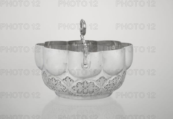 Porridge bowl porringer, Wakely & Wheeler Ltd London; goldsmiths; fl. 1791–1958, hemispherical bowl decorated with relief acanthus rosettes in the lower part, knobbed above, octave-shaped rim; two volute handles on the sides one half-preserved, rosette ornament, porridge dish, relief decoration, acanthus, 1891/1892, London, Great Britain; dish. Dimensions: height 7.6 cm, width 24 cm, depth 19.5 cm; weight 415 g., A reflective metal bowl with a scalloped rim and a curved handle rising from its center, showing a patterned band of embossed floral motifs around its lower exterior; the surface appears shiny and polished with visible reflections and some small surface marks, and the background is a smooth, light gray that fades subtly toward the edges, with the bowl casting a faint reflection on the surface beneath. The visible colors are shades of silver and gray.