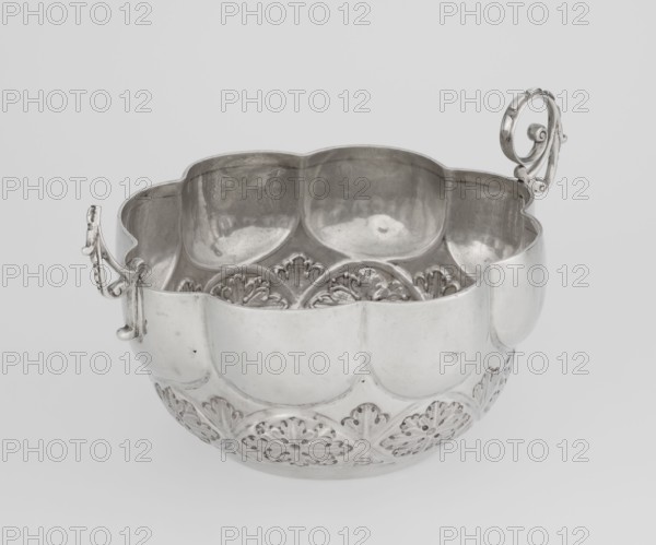 Porridge bowl porringer, Wakely & Wheeler Ltd London; goldsmiths; fl. 1791–1958 maker, porringer porridge dish with relief decoration: hemispherical bowl decorated with relief acanthus rosettes in the lower part, knobbed above, octave-shaped rim; two volute handles on the sides one half-preserved. Motifs: rosette, acanthus. Date: 1891/1892. Place: London, England, Great Britain. Materials/technique: dish; height 7.6 cm, width 24 cm, depth 19.5 cm, weight 415 g., A polished metal bowl with a scalloped rim and an overall rounded shape, shown from an angled top view revealing its interior and exterior; the exterior features a repeating decorative pattern of raised floral or fan-like motifs around the lower portion, while the upper portion has smooth, rounded panels; the interior reflects light and shows areas of subtle texture and shallow ornamental ridges near the base; two ornate handles with curled and scroll-like designs extend from opposite sides of the rim; the visible colors are si...
