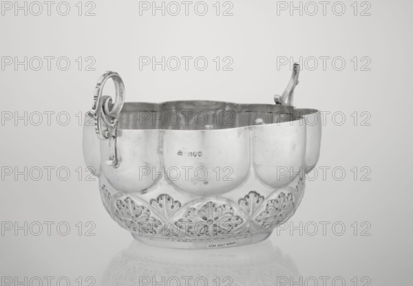Porridge bowl porringer, Wakely & Wheeler Ltd London; goldsmiths; fl. 1791–1958, hemispherical bowl decorated with relief acanthus rosettes in the lower part, knobbed above, octave-shaped rim; two volute handles on the sides one half-preserved, rosette ornament, relief decoration, porridge dish, acanthus motif, 1891/1892, London, England, Great Britain; dish. Dimensions: height 7.6 cm, width 24 cm, depth 19.5 cm; weight 415 g., A metallic decorative bowl with an ornate rim and two handles sits centered on a reflective surface; the bowl has a scalloped upper edge and a band of carved floral or shell-like motifs around its lower half, and the inside shows dents and reflective highlights; the handles have curved, scroll-like shapes with ornamental detailing; the scene is rendered in shades of gray, silver, white, and muted black tones.
