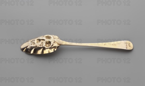 Jam spoon, by Peter, Ann & William Bateman fl. 1800–1805 and Bateman London; goldsmith's firm; fl. 1761–1843. Jam spoon with an ovoid, serrated scoop, decorated with a bunch of repoussé fruit; widening handle with a semicircular tip, slightly curved upwards, decorated with an engraved ornament of leaves and snake-like lines; on the handle in a heart-shaped medallion, engraved with a tied monogram MEK. 1803–1804, London, England. Cutlery. Height 20.7 cm; width 4.2 cm., A single metallic spoon rests on a flat surface, shown horizontally with the bowl to the left and the handle extending to the right; the spoon has a reflective gold color with darker gold highlights and small areas of brownish reflections, and the surface of the bowl features an ornate, embossed floral and shell-like pattern with raised shapes and scalloped edges, while the handle is smooth and tapers slightly with a rounded end, showing a few subtle scratches and polished sheen against a neutral gray background.