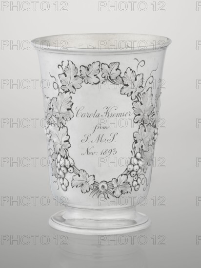 Conical silver mug on a circular foot by Barnard, Edward & Sons Ltd London; goldsmiths; fl. ca. 1680–, slightly flared profiled rim, gilded interior. Belly decorated with relief vine wreaths; one wreath bears an English inscription: Carola Kremser / from / SMS/ Nov: 1893. Date noted 1861/1862, England; height 12 cm, diameter 9 cm, weight 175 g., The image shows a metallic cup with an outward flaring rim and a rounded pedestal base, rendered in shades of silver and gray. The cup surface features an ornate raised garland of leaves and clusters of berries wrapping around the front, with textured leaf veins and curled tendrils. The interior and rim appear slightly darker silver, while the exterior is a polished light silver. The background is a smooth gradient of pale gray.