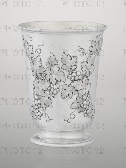 Conical mug, silver on circular foot; belly decorated with relief vine wreaths, one with an English inscription: Carola Kremser / from / SMS/ Nov: 1893. Slightly flared, profiled rim. Gilded interior. Maker: Barnard, Edward & Sons Ltd London, goldsmith's firm, active ca. 1680–. Date: 1861/1862. Place: London, England. Function: vessel. Height 12 cm; diameter 9 cm; weight 175 g. Provenance: scrap - purchase, purchase; dedications; relief decoration; vine ornament., A metal cup with a flared rim and rounded foot, finished in a reflective silver tone, decorated with an engraved pattern of vine leaves, curling tendrils, and clusters of round berries rendered in darker engraved lines and shaded areas; the surface shows subtle variations in sheen and faint wear marks, and the background is a smooth neutral gray-white.