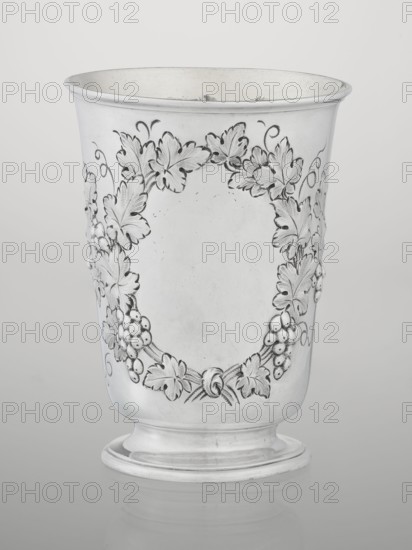 Conical mug by Edward Barnard & Sons Ltd, English goldsmiths fl. ca. 1680–, silver conical mug on a circular foot with slightly flared, profiled rim and gilded interior. Belly decorated with relief vine wreaths; one wreath contains an English inscription: Carola Kremser / from / SMS/ Nov: 1893. Dated 1861/1862, London, England. Vessel; height 12 cm, diameter 9 cm, weight 175 g., A single ornate metal cup displayed upright on a reflective surface, with an outward flaring rim and a stepped round base. The cup has a raised decorative wreath motif wrapping around its body made of stylized leaves and clusters of rounded berries, with engraved textures and subtle shadowing that emphasize the relief. The metal surface appears highly polished with visible slight scratches and small areas of darker patina in recessed details. The overall colors visible are shades of silver and gray with darker gray accents in the engraved and recessed areas, and a neutral light gray background.