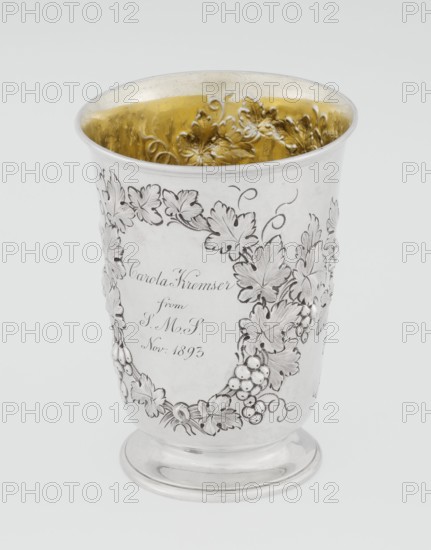Conical mug by Barnard, Edward & Sons Ltd London, goldsmith's firm, active ca. 1680– ; silver conical mug on a circular foot; belly decorated with relief vine wreaths, one with an English inscription: Carola Kremser / from / SMS / Nov: 1893. Slightly flared, profiled rim. Gilded interior. 1861/1862. England. Height 12 cm; diameter 9 cm; weight 175 g., A metallic cup with a reflective silver exterior and a gold-toned interior, decorated with an embossed wreath of leaves and grape clusters encircling its surface, sitting on a rounded silver base; the leaves and grapes are raised and detailed, and the inner rim shows textured floral embossing in the gold area.